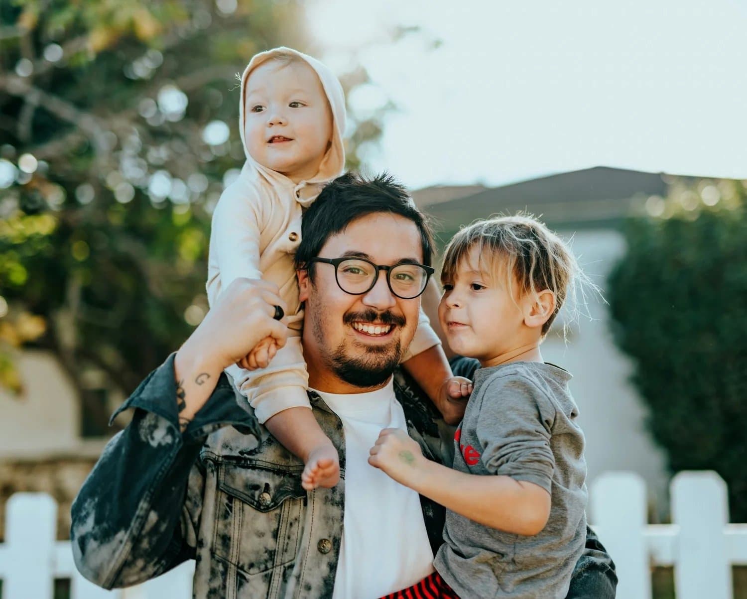 A smiling father carrying his two young children outdoors.
